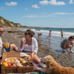 Create a realistic editorial lifestyle image of a restorative summer picnic at Seaham Glass Beach on the North Sea, showing a happy couple relaxing on a beach blanket beside a woven picnic basket filled with pasta, mangoes, sweets, juice, eggs, water, and chocolates. One person is crouched near the shoreline searching for sparkling transparent sea-glass stones, while children play, dogs run, and gentle waves roll in beneath a vivid blue sky with a few soft clouds. Capture a warm, candid, family-friendly long-weekend atmosphere after a draining workweek, with natural sunlight, crisp coastal details, subtle glass-stone sparkle, polished magazine-style realism, and no text, logos, or branding.